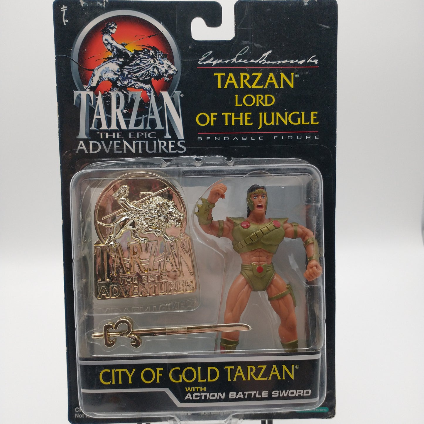 The front of the cart and bubble. The action figure is inside wearing a gold chestplate, gold underwear, and a gold headband. To the left of the action figure is a gold chrome medallion, and a gold chrome sword.